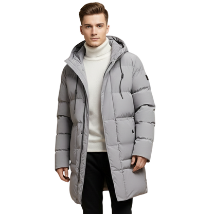 Mężczyzna ubrany w kurtkę Arkadya Outlet Kurtka Parka Męska Długa Pikowana z Kapturem Zimowa Ochronna na białym golfie, czarne spodnie i szare trampki idzie pewnie na prostym tle.