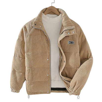 Kurtka zimowa męska strukturalny krój z czystymi liniami marki Arkadya Outlet to beżowa sztruksowa kurtka puchowa z guzikami zatrzaskowymi i wysokim kołnierzem, idealna do miejskich stylizacji, prezentowana na wieszaku na jasnym swetrze.