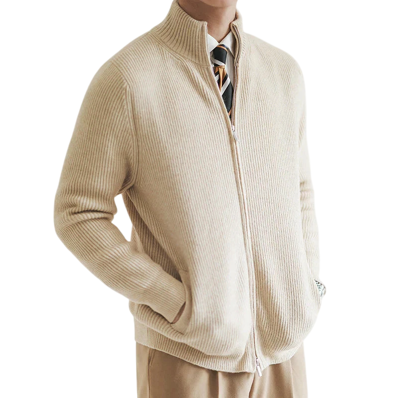 Mężczyzna stoi z rękami w kieszeniach na prostym, jasnym tle, ubrany w kurtkę Arkadya Outlet męski zapinany na zamek błyskawiczny dopasowany krój w kolorze beżowym, brązowe spodnie, białą koszulę i krawat w paski.