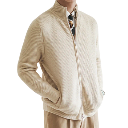 Mężczyzna stoi z rękami w kieszeniach na prostym, jasnym tle, ubrany w kurtkę Arkadya Outlet męski zapinany na zamek błyskawiczny dopasowany krój w kolorze beżowym, brązowe spodnie, białą koszulę i krawat w paski.