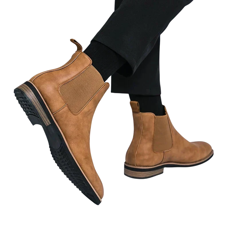 Osoba ubrana w brązowe Botki Chelsea męskie zimowe styl Chelsea na chłód by Arkadya Outlet z czarnymi elastycznymi panelami bocznymi, czarnymi spodniami, niskim obcasem i teksturowanymi gumowymi podeszwami. Tło jest w jednolitym jasnym kolorze.