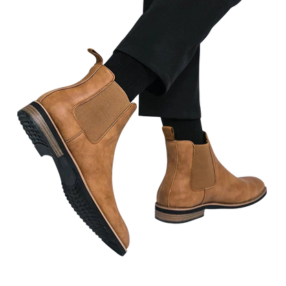 Osoba ubrana w brązowe Botki Chelsea męskie zimowe styl Chelsea na chłód by Arkadya Outlet z czarnymi elastycznymi panelami bocznymi, czarnymi spodniami, niskim obcasem i teksturowanymi gumowymi podeszwami. Tło jest w jednolitym jasnym kolorze.