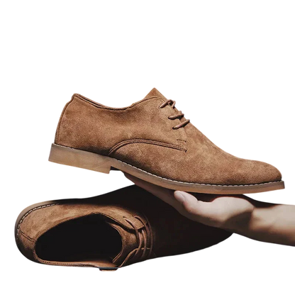 Ręka trzyma elegancki Arkadya Outlet Buty Biznesowe Męskie Płaski Obcas Zamknięty Nosek nad pasującym butem leżącym płasko na białym tle.