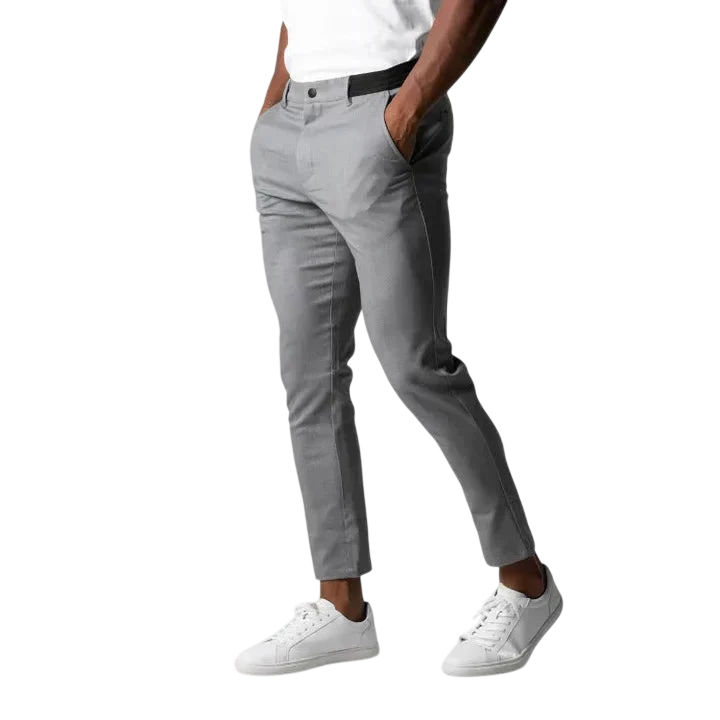 Osoba ubrana w Arkadya Outlet Spodnie chinosy męskie modny krój pełna długość użytkowy pas w kolorze jasnoszarym, biały t-shirt i białe trampki stoi z rękami w kieszeniach na jednolitym jasnoszarym tle. Głowa nie jest widoczna.