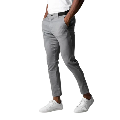 Osoba ubrana w Arkadya Outlet Spodnie chinosy męskie modny krój pełna długość użytkowy pas w kolorze jasnoszarym, biały t-shirt i białe trampki stoi z rękami w kieszeniach na jednolitym jasnoszarym tle. Głowa nie jest widoczna.