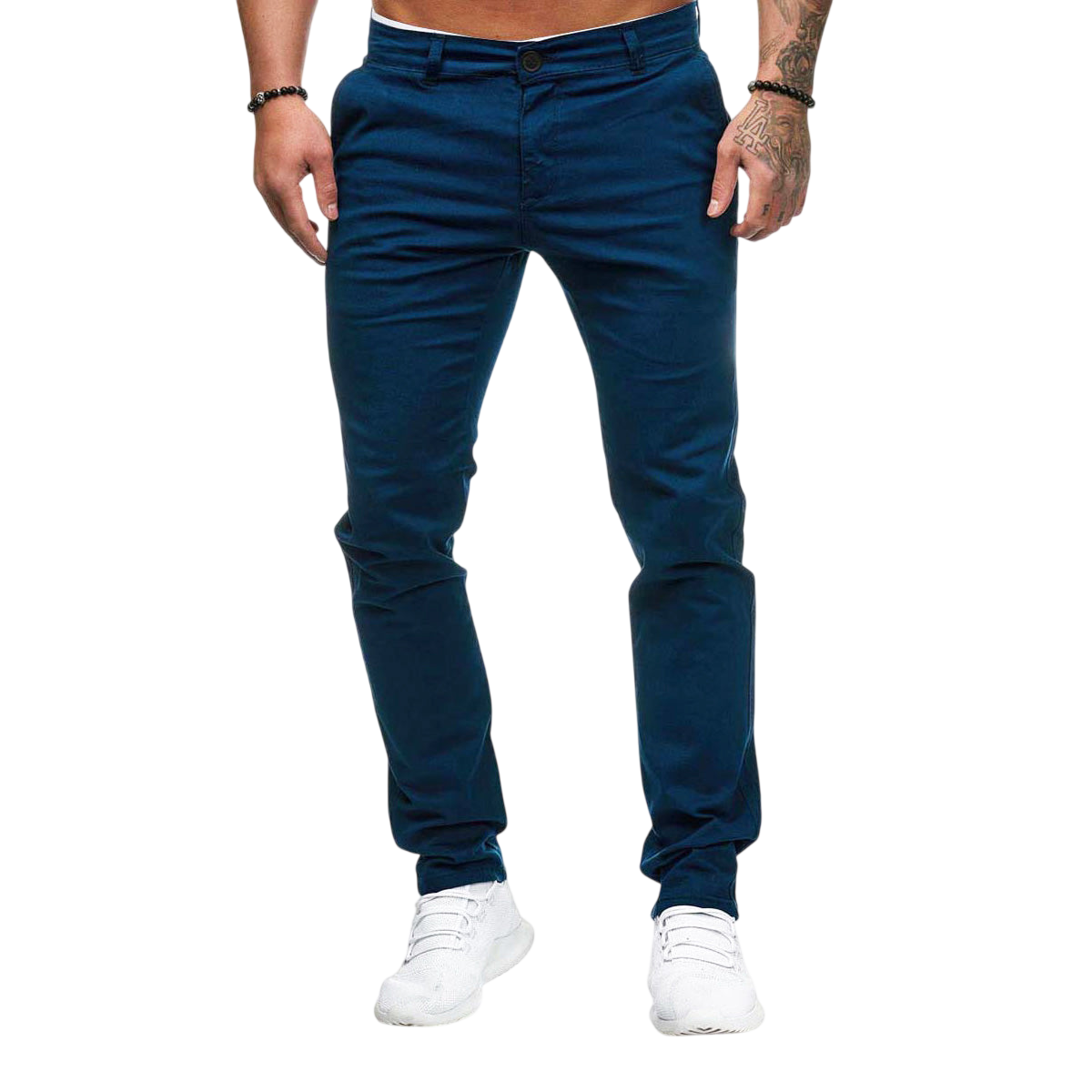 Mężczyzna modeluje Męskie spodnie Arkadya Outlet o dopasowanym kroju i prostej nogawce w kolorze niebieskim, w połączeniu z białymi sneakersami. Stoi na jasnoszarym tle z rękami w kieszeniach i tatuażami widocznymi na jednym ramieniu. Jego górna część ciała nie jest pokazana.