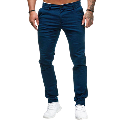 Mężczyzna modeluje Męskie spodnie Arkadya Outlet o dopasowanym kroju i prostej nogawce w kolorze niebieskim, w połączeniu z białymi sneakersami. Stoi na jasnoszarym tle z rękami w kieszeniach i tatuażami widocznymi na jednym ramieniu. Jego górna część ciała nie jest pokazana.