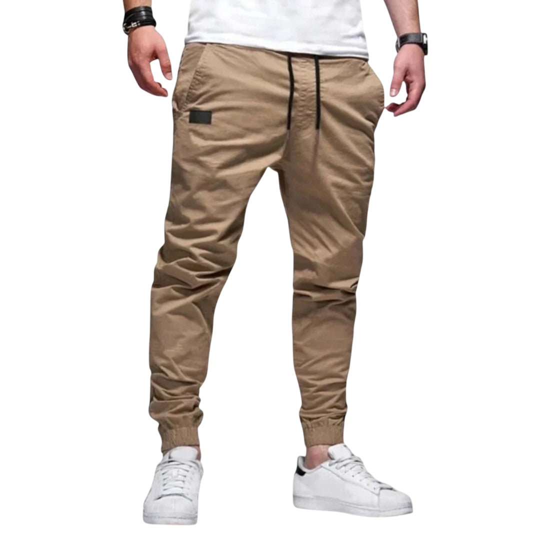 Mężczyzna w Arkadya Outlet spodnie joggerowe męskie krój slim ze ściągaczem w pasie i mankietach, biały t-shirt i białe sneakersy stoi na jednolitym jasnoszarym tle.