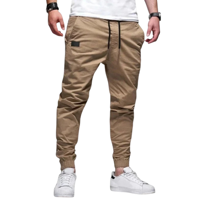 Mężczyzna w Arkadya Outlet spodnie joggerowe męskie krój slim ze ściągaczem w pasie i mankietach, biały t-shirt i białe sneakersy stoi na jednolitym jasnoszarym tle.