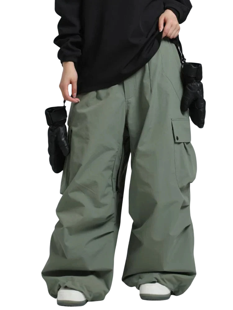 Osoba w Arkadya Outlet spodnie zimowe męskie szeroki krój regularny na warunki zimowe w szałwiowej zieleni, czarnej bluzce z długim rękawem i białych tenisówkach stoi na jednolitym tle, z czarnymi rękawiczkami zwisającymi z kieszeni spodni.