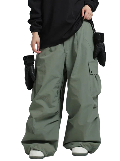 Osoba w Arkadya Outlet spodnie zimowe męskie szeroki krój regularny na warunki zimowe w szałwiowej zieleni, czarnej bluzce z długim rękawem i białych tenisówkach stoi na jednolitym tle, z czarnymi rękawiczkami zwisającymi z kieszeni spodni.