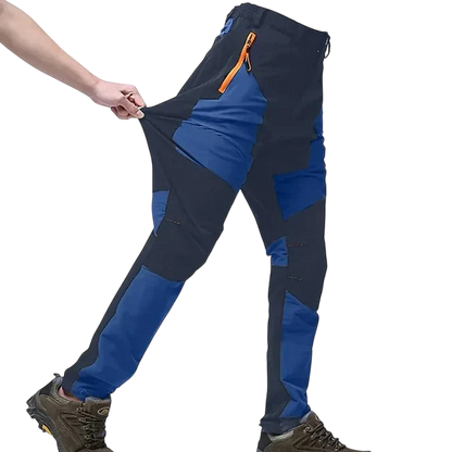 Osoba w Arkadach Outlet Spodnie Terenowe Męskie Wodoodporne Wiatroszczelne Regulowane (niebiesko-czarne, pomarańczowe kieszenie na suwak) i brązowe buty turystyczne rozciągają się, aby pokazać elastyczność - idealne do aktywności na świeżym powietrzu.