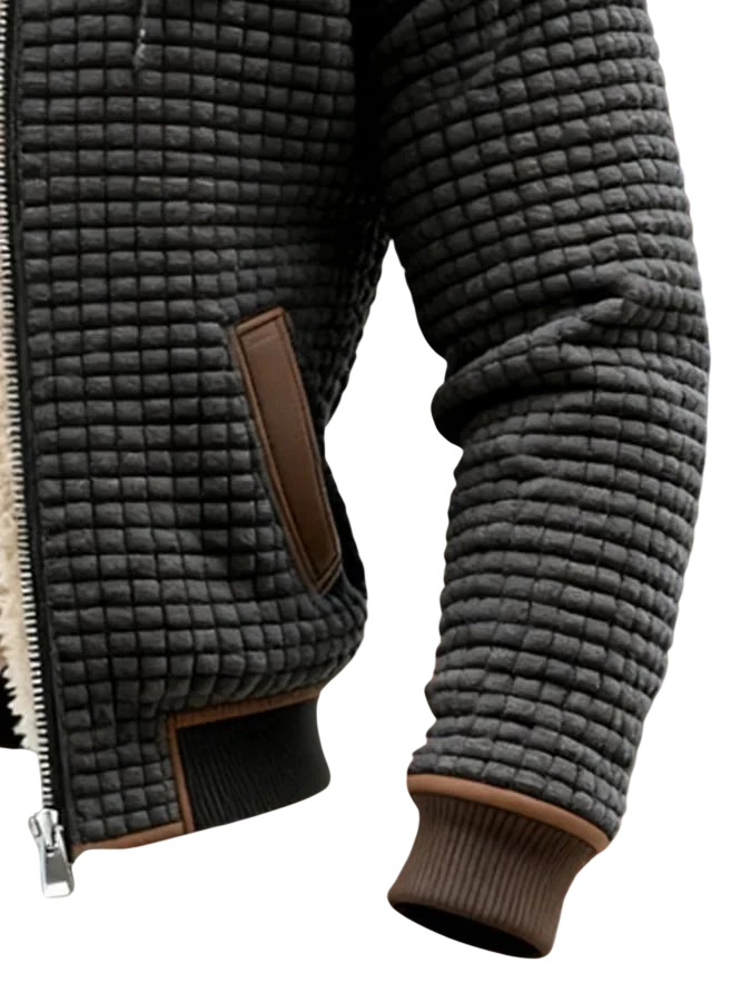 Zbliżenie osoby noszącej kurtkę Arkadya Outlet Lekka Kurtka Pikowana Męska Luźny Krój Wysoki Kołnierz, z teksturowanym ciemnoszarym wzorem w kratkę, brązowymi akcentami, zamkiem błyskawicznym i prążkowanymi mankietami, na rozmytym brązowo-zielonym tle.