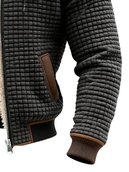 Zbliżenie osoby noszącej kurtkę Arkadya Outlet Lekka Kurtka Pikowana Męska Luźny Krój Wysoki Kołnierz, z teksturowanym ciemnoszarym wzorem w kratkę, brązowymi akcentami, zamkiem błyskawicznym i prążkowanymi mankietami, na rozmytym brązowo-zielonym tle.