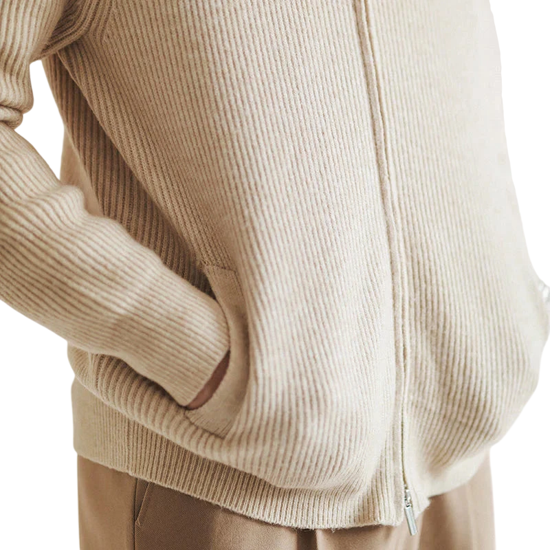 Osoba ubrana w kardigan Arkadya Outlet Męski, beżowy prążkowany sweter zapinany na zamek o dopasowanym kroju, z rękami w kieszeniach, w połączeniu z jasnobrązowymi spodniami.