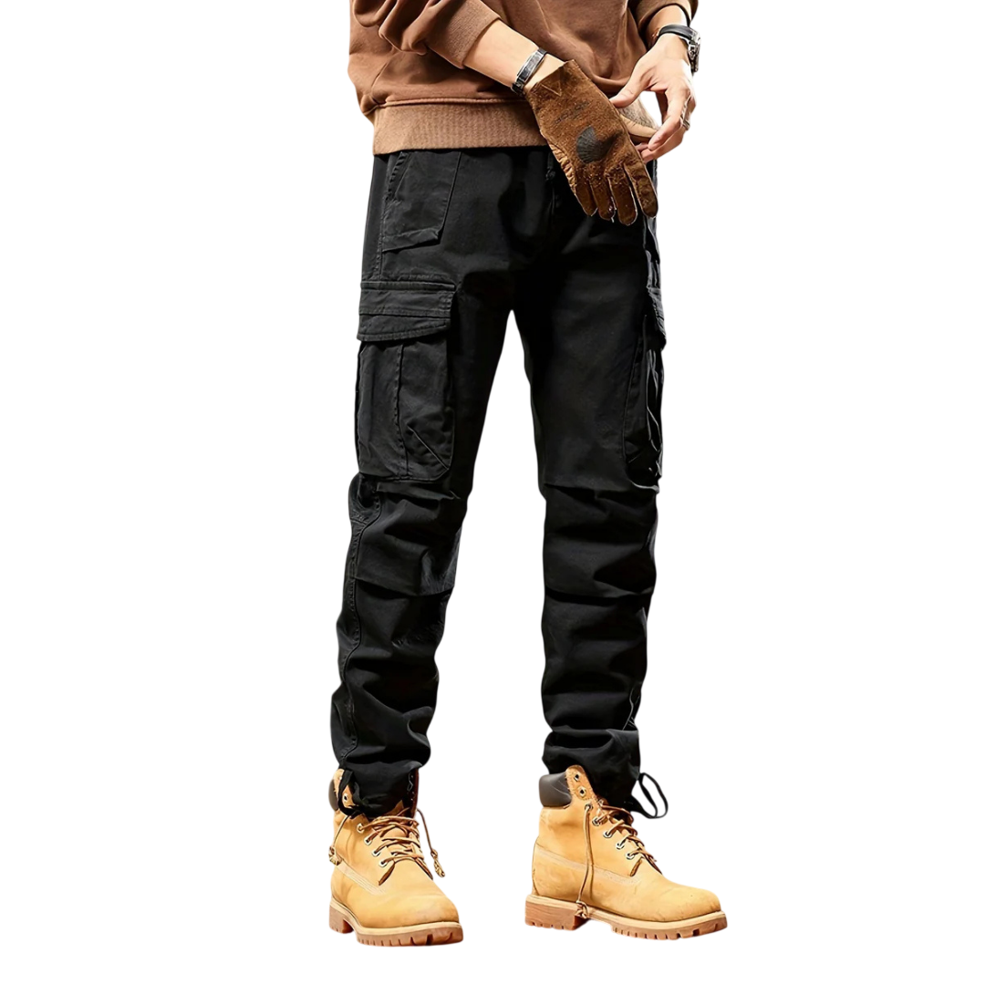 Mężczyzna ubrany w Arkadya Outlet Spodnie cargo męskie taktyczne wielokieszeniowa konstrukcja w kolorze czarnym, z brązową bluzą, brązowymi rękawiczkami i brązowymi butami roboczymi stoi na jasnym tle. Widoczna jest tylko jego dolna połowa.