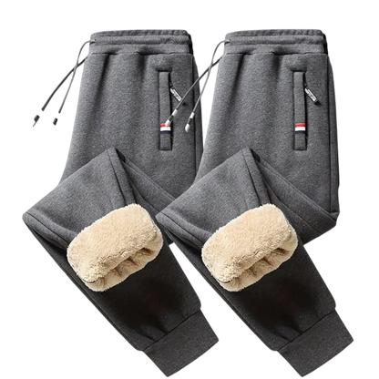 Dwie pary Arkadya Outlet Spodnie Termiczne Męskie Elastyczny Pas Z Regulacją W Talii w kolorze ciemnoszarym są pokazane na jasnej powierzchni, z zewnętrznymi kieszeniami, sznurkami i beżową polarową podszewką - idealne na chłodne dni.
