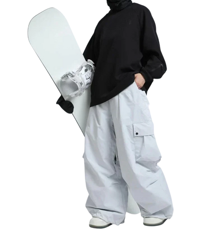 Osoba w odblaskowych goglach narciarskich, czarnym topie i Arkadya Outlet spodnie zimowe męskie szeroki krój regularny na warunki zimowe trzyma białą deskę snowboardową na jednolitym tle.