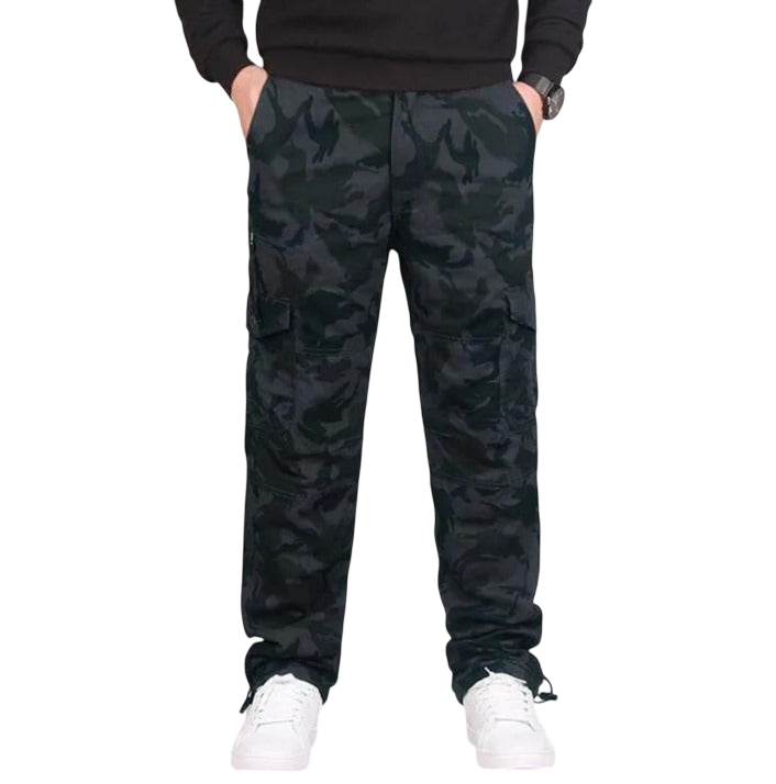 Mężczyzna ubrany w Arkadya Outlet Spodnie cargo męskie prosta nogawka z kieszeniami i zamkiem, czarną koszulę z długim rękawem i białe trampki stoi z rękami w kieszeniach na jednolitym tle.