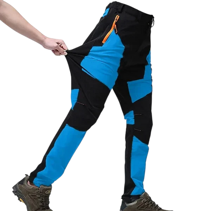 Osoba ubrana w Arkadya Outlet Spodnie Terenowe Męskie Wodoodporne Wiatroszczelne Regulowane i brązowe buty turystyczne rozciąga bok spodni, aby zademonstrować ich elastyczność na zwykłym białym tle.