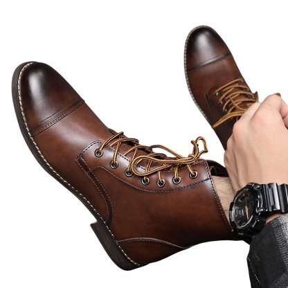 Mężczyzna w Arkadii Outlet Buty męskie wysoka cholewka z wiązaniem pełna wysokość i czarny zegarek siedzi na szarej podłodze wyłożonej kafelkami, jedna noga w butach skrzyżowana na drugiej.