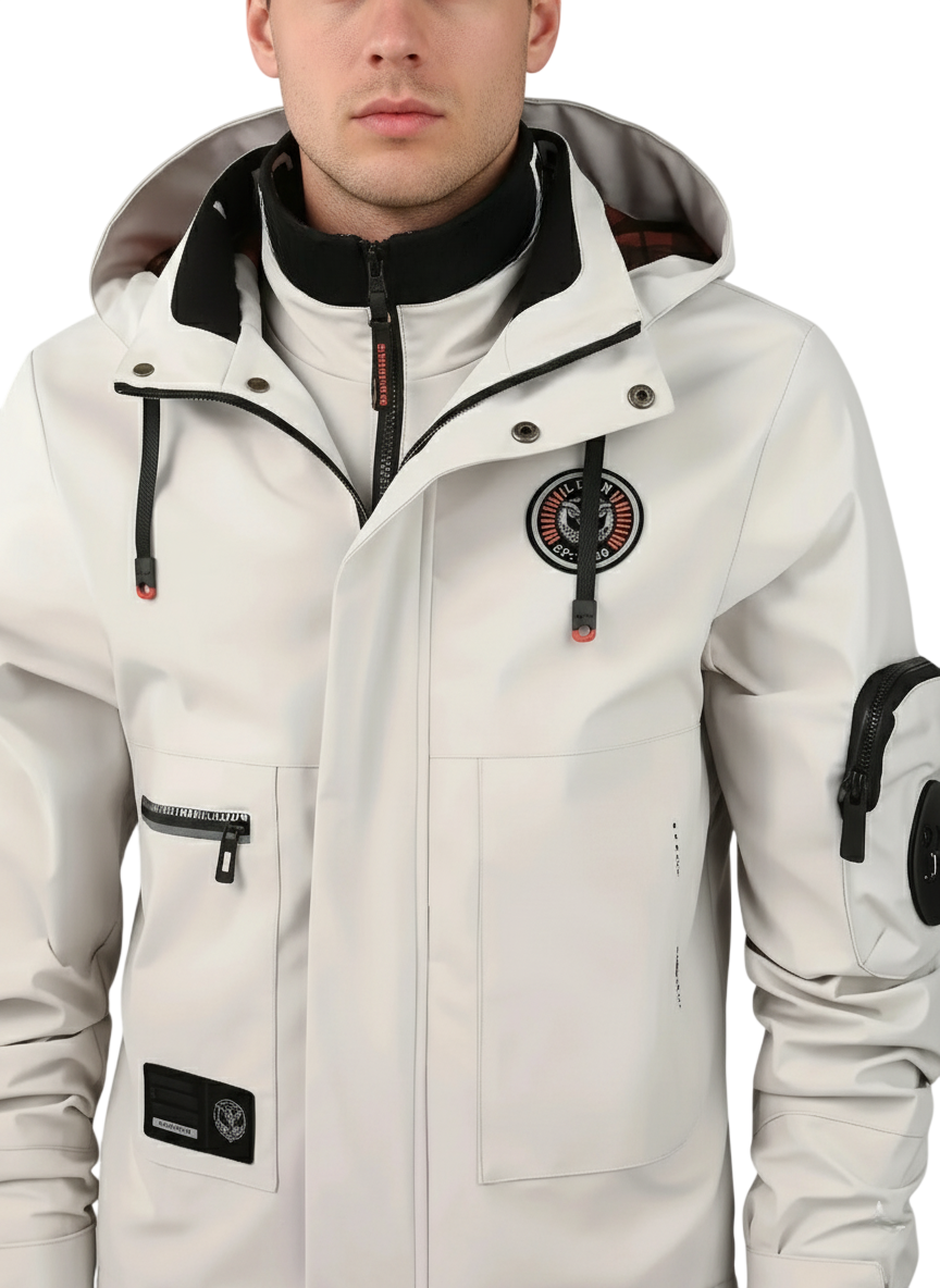 Mężczyzna ubrany w Kurtkę Zewnętrzną Męską Wodoodporną Zapięcie na Zamek by Arkadya Outlet, jasną kurtkę z czarnymi suwakami, wysokim kołnierzem i wieloma naszywkami na piersi i ramionach, pokazaną od ramion do talii na jednolitym tle.