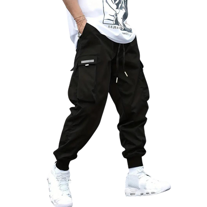 Osoba, widoczna od pasa w dół, nosi spodnie cargo Arkadya Outlet Męskie z wąską nogawką i wieloma kieszeniami, w połączeniu z białą koszulką z grafiką i białymi trampkami, stojąc na prostym szarym tle.