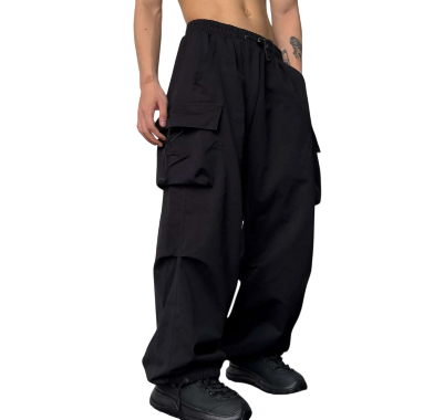 Postać stoi na tle białej ściany, pokazując tylko dolną część tułowia i nogi, ubrana w odzież męską Arkadya Outlet Męskie Spodnie z szeroka nogawką i wieloma kieszeniami, w połączeniu z czarnymi butami.