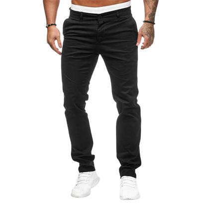 Mężczyzna ma na sobie Arkadya Outlet Męskie spodnie | Dopasowany krój prosta nogawka, białą koszulę, białe trampki, bransoletki i ujawnia tatuaże na lewym ramieniu, stojąc na prostym jasnoszarym tle.