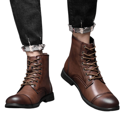 Osoba ubrana w Arkadya Outlet Buty męskie wysoka cholewka z wiązaniem pełna wysokość, brązowe skórzane sznurowane buty z czarnymi podeszwami, sparowane z czarnymi podartymi dżinsami, stojąca na podłodze wyłożonej kafelkami na tle szarej ściany.