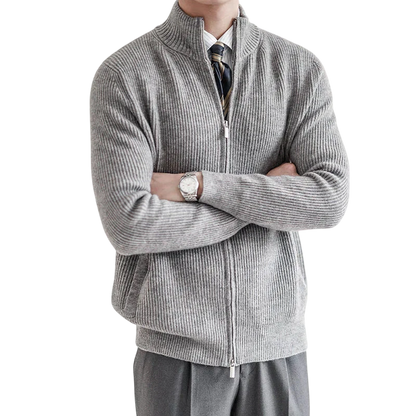 Mężczyzna stoi ze skrzyżowanymi rękami w jasnoszarym Arkadya Outlet Męski kardigan, zapinany na zamek błyskawiczny i o dopasowanym kroju. Ubrany w białą koszulę, krawat w paski i szare spodnie; twarz zasłonięta. Tło neutralne, delikatnie oświetlone.