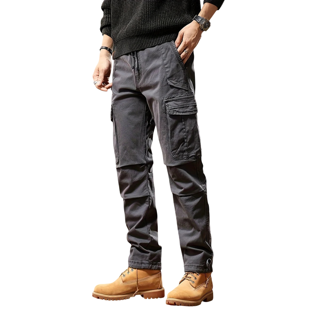 Mężczyzna w ciemnoszarym Arkadya Outlet Spodnie cargo męskie taktyczne wielokieszeniowa konstrukcja, czarnym swetrze, zegarku i brązowych sznurowanych butach roboczych stoi z jedną ręką w kieszeni na prostym białym tle.