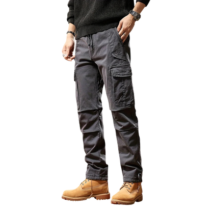 Mężczyzna w ciemnoszarym Arkadya Outlet Spodnie cargo męskie taktyczne wielokieszeniowa konstrukcja, czarnym swetrze, zegarku i brązowych sznurowanych butach roboczych stoi z jedną ręką w kieszeni na prostym białym tle.