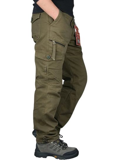 Osoba ma na sobie Arkadya Outlet Spodnie cargo męskie prosta nogawka z kieszeniami i zamkiem oraz szare buty turystyczne, stoi z jedną ręką w kieszeni. Spodnie mają wiele kieszeni, zamki błyskawiczne, metkę na szlufce, na białym tle.
