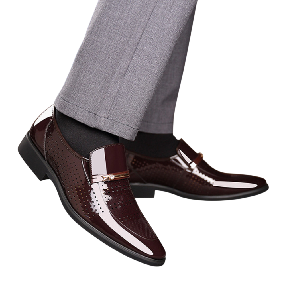 Osoba nosi błyszczące ciemnobrązowe Mokasyny Wizytowe Męskie Wsuwany Szpiczasty Nosek by Arkadya Outlet, z błyszczącym wykończeniem i metalicznymi detalami, w połączeniu z czarnymi skarpetkami i jasnoszarymi spodniami.