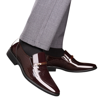 Osoba nosi błyszczące ciemnobrązowe Mokasyny Wizytowe Męskie Wsuwany Szpiczasty Nosek by Arkadya Outlet, z błyszczącym wykończeniem i metalicznymi detalami, w połączeniu z czarnymi skarpetkami i jasnoszarymi spodniami.