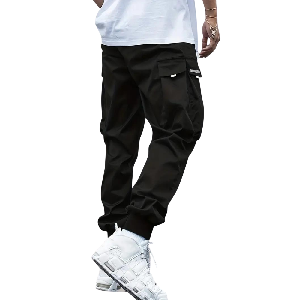 Osoba ma na sobie Arkadya Outlet Męskie spodnie cargo | Dopasowana nogawka z wieloma kieszeniami, w połączeniu z białą koszulką, skarpetkami i trampkami, stojąca na jednolitym jasnoszarym tle. Górna część ciała jest częściowo poza kadrem.