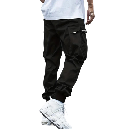 Osoba ma na sobie Arkadya Outlet Męskie spodnie cargo | Dopasowana nogawka z wieloma kieszeniami, w połączeniu z białą koszulką, skarpetkami i trampkami, stojąca na jednolitym jasnoszarym tle. Górna część ciała jest częściowo poza kadrem.