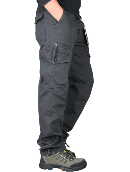 Mężczyzna ubrany w Arkadya Outlet Spodnie cargo męskie prosta nogawka z kieszeniami i zamkiem oraz szare buty turystyczne stoi bokiem z jedną ręką w kieszeni na jasnym tle.