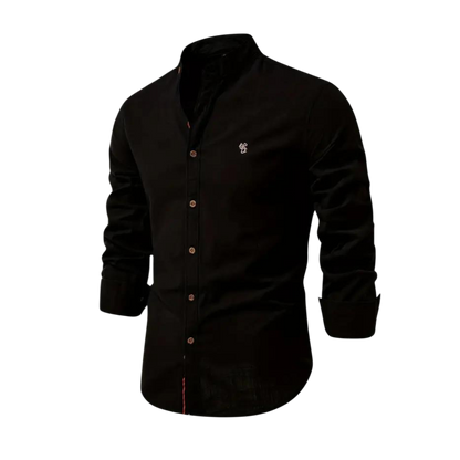 Koszula męska Arkadya Outlet z kołnierzem stójka, w czerni, długi rękaw, krój regularny, małe białe logo na lewej piersi. Idealna do codziennego użytku. Prezentowana na tle jasnego, rozmytego wnętrza.