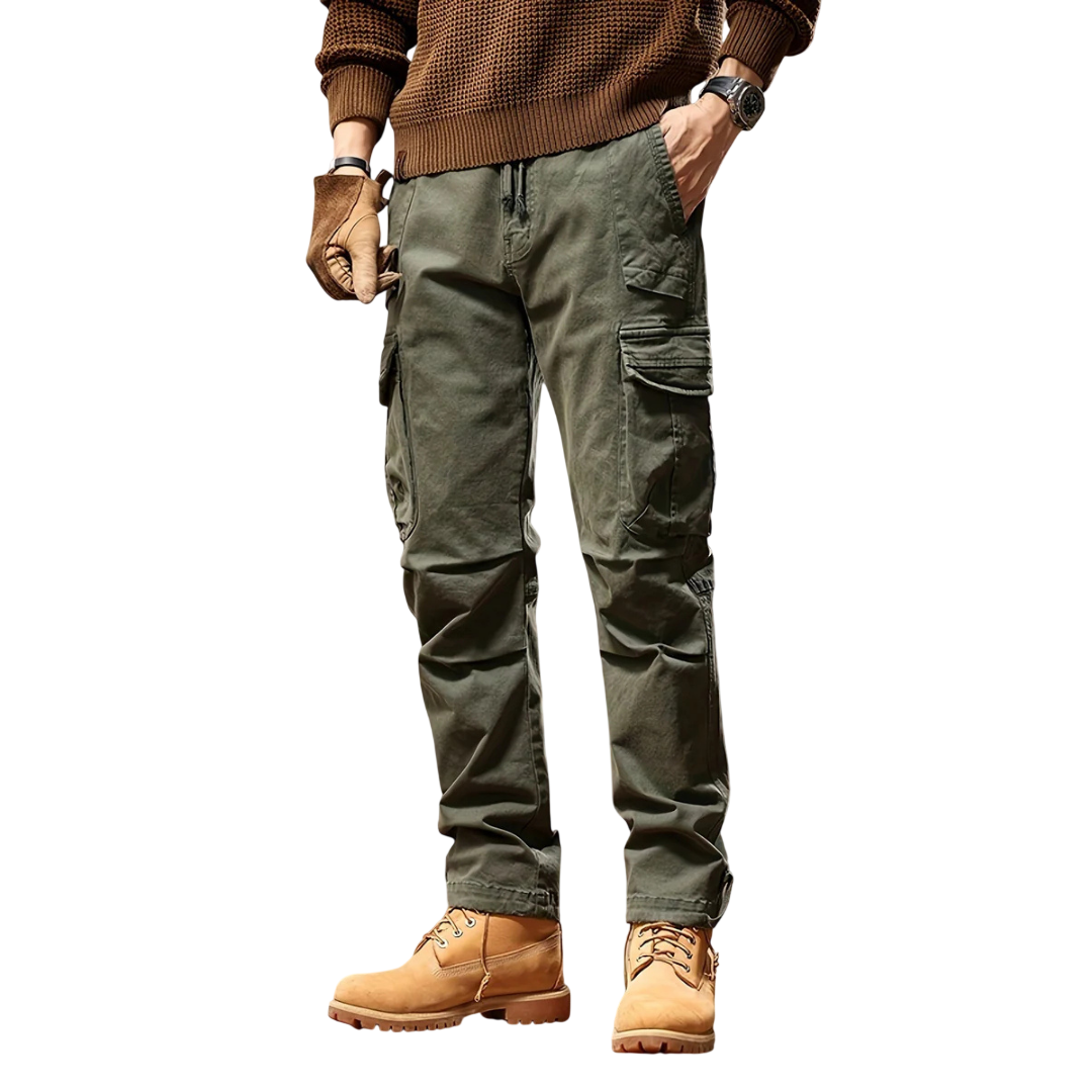 Osoba modeluje Arkadya Outlet Spodnie cargo męskie taktyczne wielokieszeniowa konstrukcja w kolorze oliwkowym, w połączeniu z brązowym swetrem, rękawiczkami, zegarkiem na rękę i brązowymi butami roboczymi. Stoi na białym tle z rękami w kieszeniach; twarz niewidoczna.