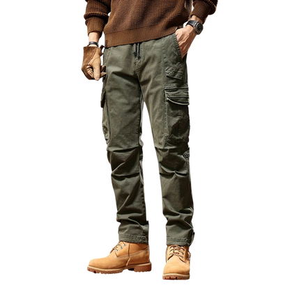 Osoba modeluje Arkadya Outlet Spodnie cargo męskie taktyczne wielokieszeniowa konstrukcja w kolorze oliwkowym, w połączeniu z brązowym swetrem, rękawiczkami, zegarkiem na rękę i brązowymi butami roboczymi. Stoi na białym tle z rękami w kieszeniach; twarz niewidoczna.