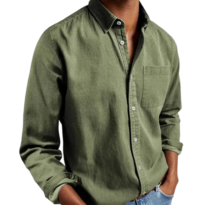 Osoba modeluje Arkadya Outlet Koszula męska klasyczny krój prosta linia regularna długość w kolorze oliwkowym, podwinięte rękawy, w połączeniu z jasnoniebieskimi dżinsami i rękami w kieszeniach - elegancki styl koszuli do pracy.