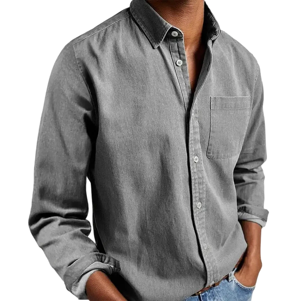 Osoba nosi Arkadya Outlet Koszula męska klasyczny krój prosta linia regularna długość w kolorze szarym z kieszenią na piersi i niebieskimi dżinsami, stojąc z rękami w kieszeniach na jednolitym jasnoszarym tle - idealna koszula do pracy.