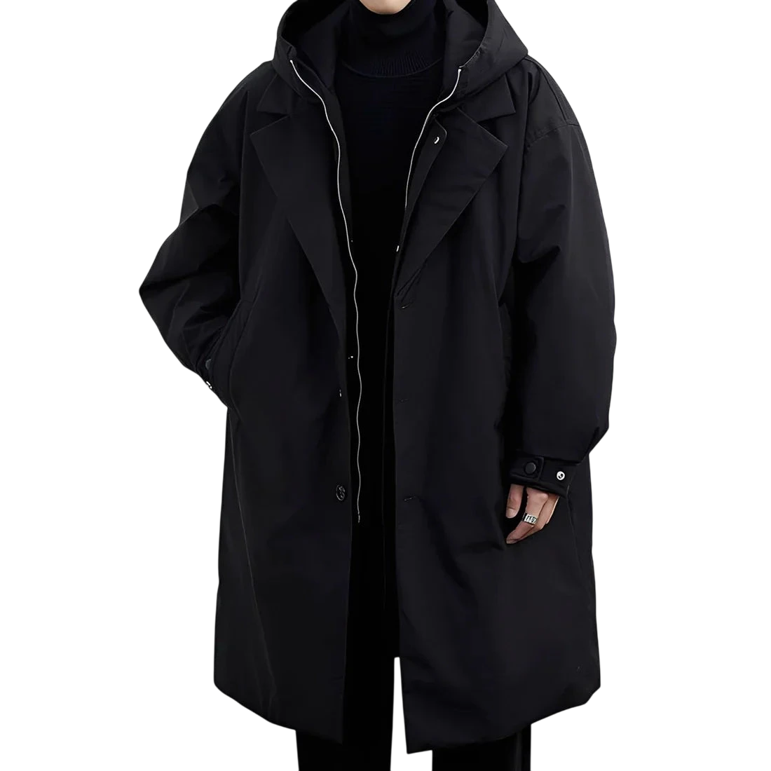 Osoba stoi na białym tle ubrana w czarne spodnie, czarny golf i Arkadya Outlet Płaszcz męski wodoodporny długi krój z kapturem podszewką, płaszcz oversize z rękami schowanymi w kieszeniach.
