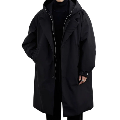 Osoba stoi na białym tle ubrana w czarne spodnie, czarny golf i Arkadya Outlet Płaszcz męski wodoodporny długi krój z kapturem podszewką, płaszcz oversize z rękami schowanymi w kieszeniach.